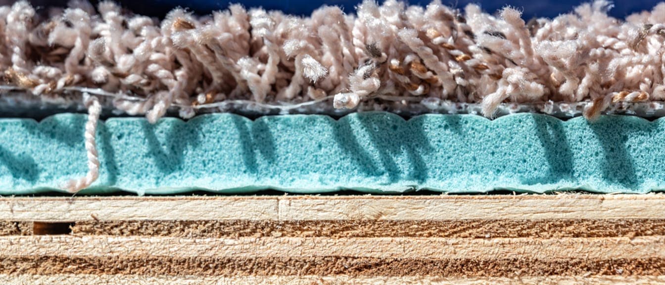 Cross-section of a carpeted floor showing the pile on top, the foam pad beneath it, and the plywood subfloor at the bottom